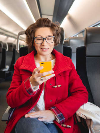 Smiling woman in red duffle coat texting on smartphone in suburban train. Travel by land vehicle.の写真素材
