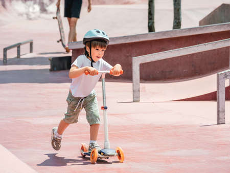 Little boy rides kick scooter in skate park. Special concrete bowl structures in urban park. Training to skate at summer.の写真素材