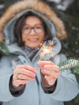 Smiling woman celebrates Christmas or New Year with bright sparkler. Bengal fire, traditional firework for winter holiday celebration outdoors.の写真素材