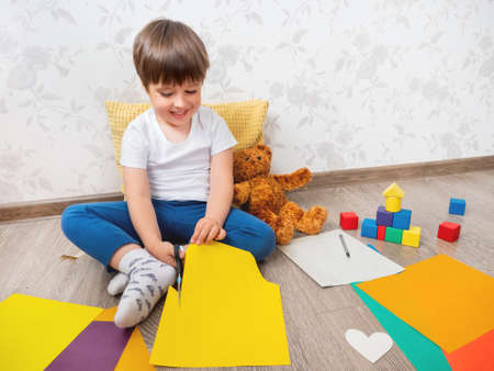 Toddler boy learns to cut colored paper with scissors. Kid sits on floor in kids room with toy blocks and teddy bear. Educational classes for children. Developing feeling sensations and fine motor skills at home.の写真素材
