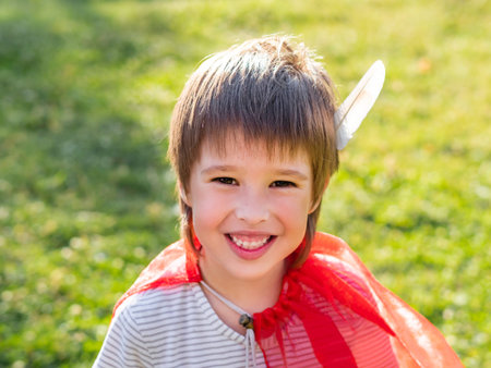 Portrait of smiling boy playing American Indian. Kid with white bird feather and red cloak. costume role play. outdoor leisure activities.の写真素材