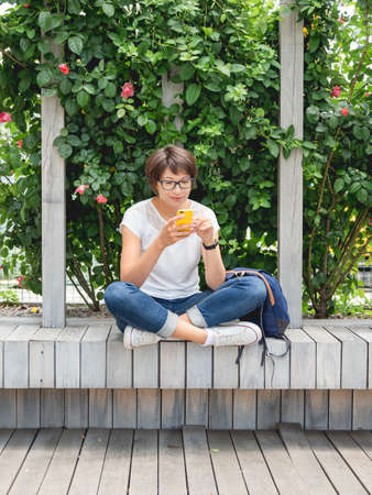 Woman is texting on smartphone. Pretty woman is sitting with smartphone on wooden bench in public park. summer vibes.の写真素材