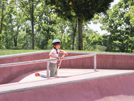 Little boy rides kick scooter in skate park. Special concrete bowl structures in urban park. Training to skate at summer.の写真素材
