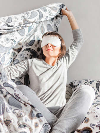 Young woman in gray pajama and sleeping mask on face. She is just woke up and sit in bed. Early morning in cozy home.の写真素材