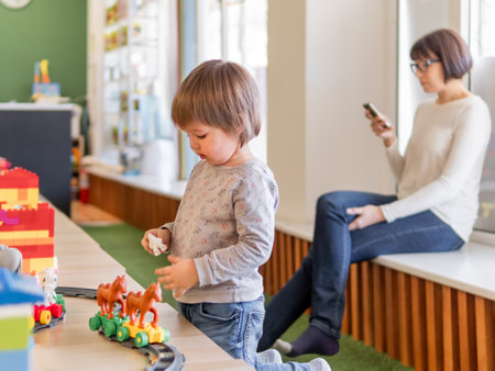 Toddler plays with colorful toy blocks while his mother or babysitter texting in smartphone. Little boy stares on toy constructor. Interior of kindergarten or nursery.の写真素材