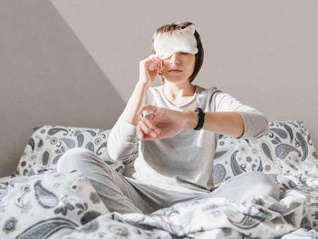 Sleepy woman in gray pajama and sleeping mask on face. She is just woke up and sit in bed. Early morning in cozy home.の写真素材