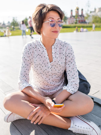Woman in colorful sunglasses sits on wooden open stage in public park. Pretty female in casual clothes texting on smartphone.の写真素材