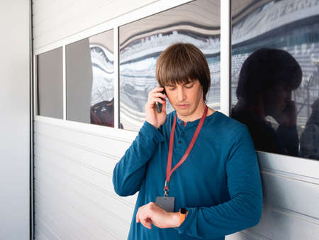 Man with badge on red ribbon talks by smartphone and looks on wristwatch on pit lane at autodrom. sports car racing. Sports competition on special track.の写真素材