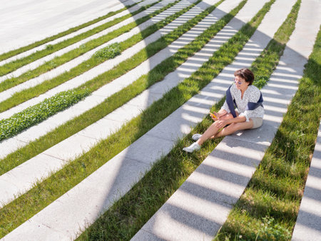 Woman texting in her smartphone while sits on bench in urban park. Striped shadow on green lawn. Modern lifestyle of millennials.の写真素材