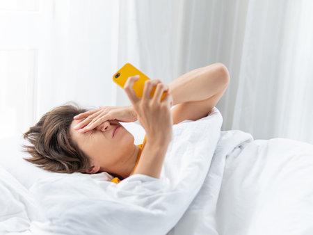 Sleepy woman looks on smartphone screen. Using cellular phone as alarm clock. Wireless technology in bed.の写真素材