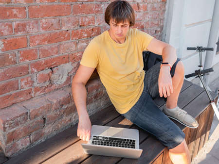 Man sits with laptop and kick scooter in street cafe. Freelancer at work. Student learns remotely from outdoors. Modern lifestyle. Summer vibes. Open air workplace.の写真素材