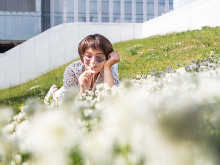 Woman in colorful sunglasses sniffs clover flowers on lawn in urban park. Nature in town. Relax outdoors after work. Summer vibes.の写真素材
