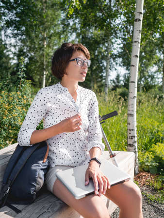 Woman works with laptop on wooden bench in urban park. Summer heat in town. Modern workplace for freelancers. Student educates online from outdoors.の写真素材