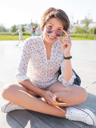 Woman in colorful sunglasses sits on wooden open stage in public park. Pretty female in casual clothes texting on smartphone.の写真素材
