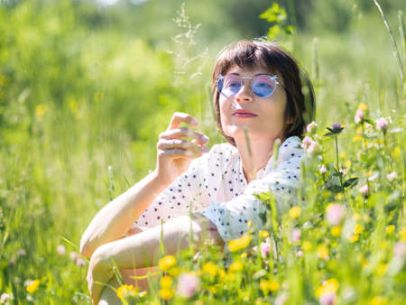 Woman in colorful sunglasses, enjoys sunlight and flower fragrance on grass field. summer vibes. Relax outdoors. Self-soothing.の写真素材