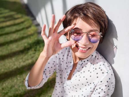 Smiling woman in colorful sunglasses has a rest on lawn in urban park. nature in town. Relax outdoors after work. summer vibes.の写真素材