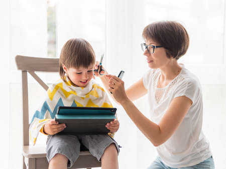 Mother cuts her son's hair by herself. Little boy sits with digital tablet. New normal in case of coronavirus COVID-19 quarantine and lockdown.の写真素材