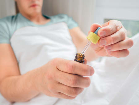 Ill man is lying in bed with liquid medicine. Man is measuring drops. Feeling sick because of disease and temporary disability.の写真素材