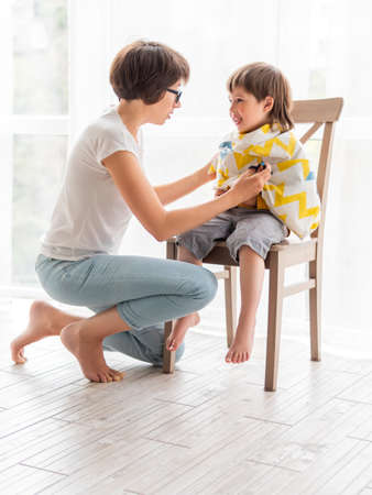 Mother cuts her son's hair by herself. Little boy sits, covered with cloth, and holds pair of scissors. family life.の写真素材