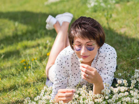 Woman in colorful sunglasses sniffs clover flowers on lawn in urban park. Nature in town. Relax outdoors after work. Summer vibes.の写真素材