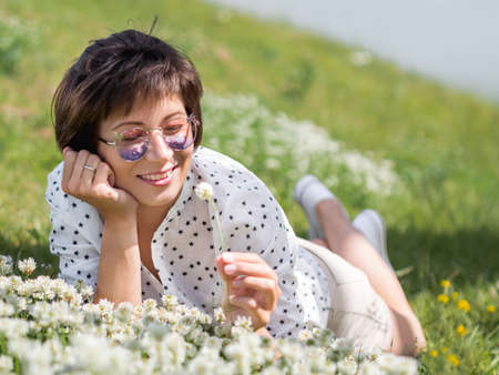 Woman in colorful sunglasses sniffs clover flowers on lawn in urban park. Nature in town. Relax outdoors after work. Summer vibes.の写真素材