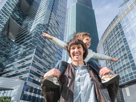 Laughing boy plays like airplane on father's shoulders. Dad and son looks on glass walls of buildings. Future and modern technologies, life balance and family life in well keeps districts.の写真素材