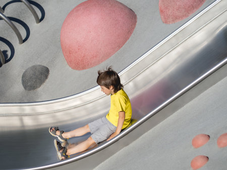 Boy with his eyes closed rides down a metal slide on modern children's sports and playground. Kid overcomes fear and learns new things. Active leisure outdoors. sport lifestyle.の写真素材