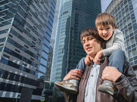 Little boy sits on father's shoulders between skyscrapers. Dad and son looks on glass walls of buildings. Future and modern technologies, life balance and family life in well keeps districts.の写真素材