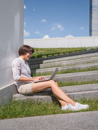 Woman sits with laptop on urban park bench. Freelancer at work. Student learns remotely from outdoors. Modern lifestyle. Summer vibes. Outdoor workplace.の写真素材