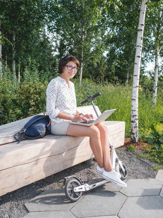 Smiling woman works with laptop on wooden bench in urban park. summer vibes. Modern workplace for freelancers. Student educates online from outdoors.の写真素材