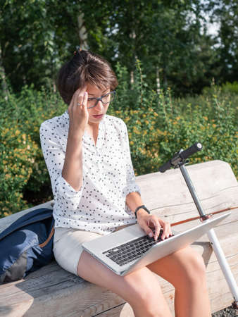 Puzzled woman works with laptop on wooden bench in urban park. summer vibes. Modern workplace for freelancers. Student educates online from outdoors.の写真素材