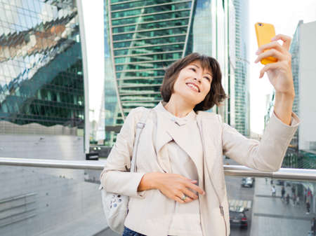 Woman with yellow smartphone takes selfie on big city background. Modern buildings with glass wall. Millennial lifestyle.の写真素材