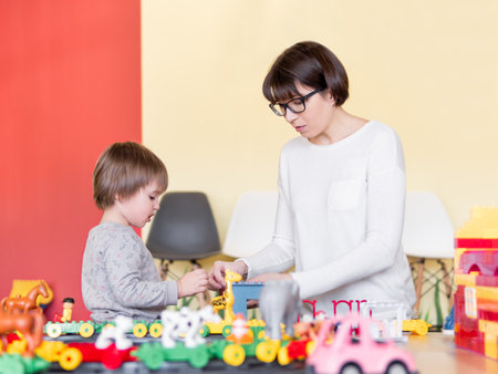 Toddler plays with colorful toy blocks while his mother or babysitter. Little boy stars on toy constructor. Interior of kindergarten or nursery.の写真素材