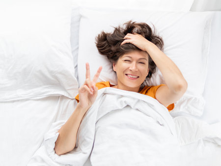Top view on sleepy woman showing Victory gesture in bed. Woman stretches while lying on white bed linen. Pleasant awakening. good sleep.の写真素材
