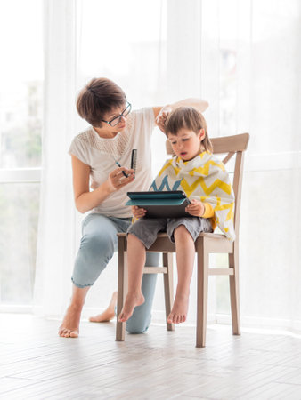 Mother cuts her son's hair by herself. Little boy sits, covered with cloth, and holds digital tablet. new normal.の写真素材