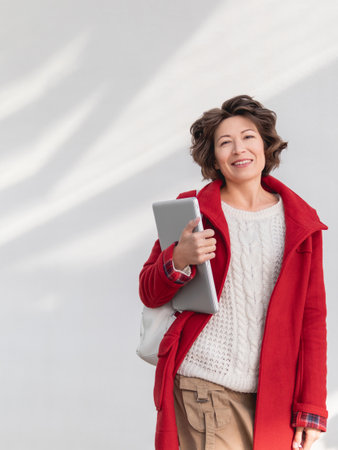 Curly woman in red coat is standing by white wall. Smiling adult student with laptop. Freelancer with portable device for work. modern lifestyle.の写真素材