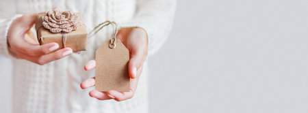 Woman in white cable knitted sweater holds Christmas present in craft paper with hand crocheted flower. Clear tag for your text. New Year mock up. Horizontal background with copy space.の写真素材