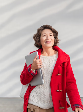 Curly woman in red coat is standing by white wall. Smiling adult student with laptop. Freelancer with portable device for work. modern lifestyle.の写真素材