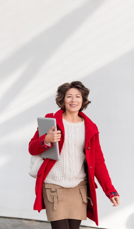Curly woman in red coat. Smiling adult student with laptop. Freelancer with portable device for work. modern lifestyle. White wall with copy space.の写真素材