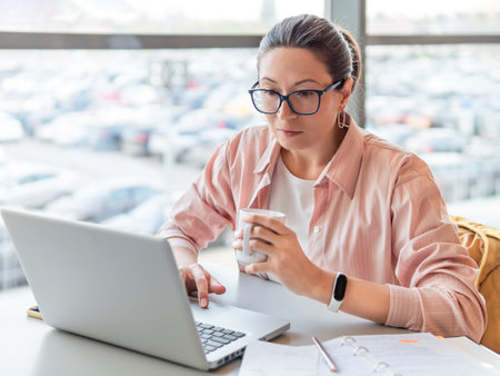 Woman in eyeglasses works with laptop. Modern office with panoramic windows at co-working center. Workplace for freelancers in business center.の写真素材
