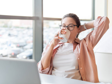 Relaxed woman drinks coffee and works with laptop. Modern office with panoramic windows at co-working center. Workplace for freelancers or students in business center.の写真素材
