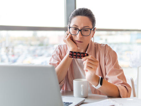 Frowning woman looks on medicine pills while working with laptop. Mental health problems, emotional burnout or headache. Modern office at co-working center. Workplace for freelancers or students.の写真素材