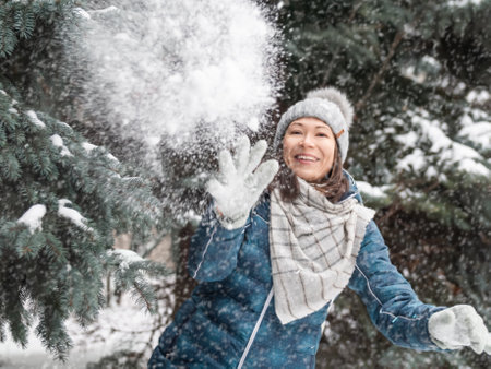 Smiling woman in cable knitted hat is playing with snow. Fun in park between snowy fir trees. Woman laughs as she is throwing a snowball. cold season. soft focus.の写真素材