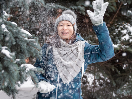 Smiling woman in cable knitted hat is playing with snow. Fun in park between snowy fir trees. Woman laughs as she is throwing a snowball. cold season.の写真素材