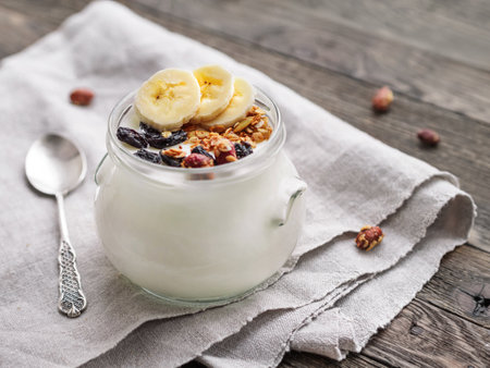 Natural homemade yogurt in a glass jar. Healthy food for breakfast with muesli. Jar with granola and banana slices on linen tablecloth on wooden table.の写真素材