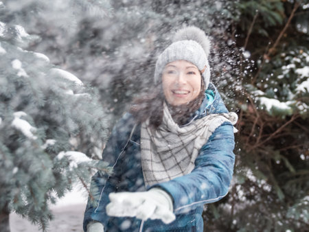 Smiling woman in cable knitted hat is playing with snow. Fun in park between snowy fir trees. Woman laughs as she is throwing a snowball. cold season.の写真素材