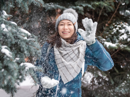Smiling woman in cable knitted hat is playing with snow. Fun in park between snowy fir trees. Woman laughs as she is throwing a snowball. cold season.の写真素材