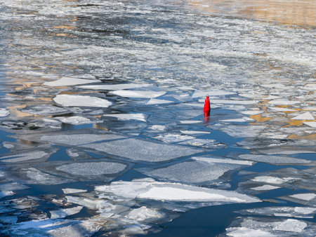 Bright red buoy among ice floes. Ice drift on river at spring day. Sunny weather at the beginning of warm season.の写真素材