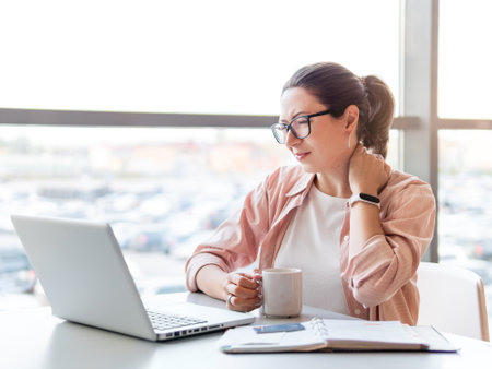 Woman winces from pain in her neck. Female office employee tries to stretch stiff neck muscles while working with laptop. Modern office at co-working center. Workplace for freelancers or students.の写真素材