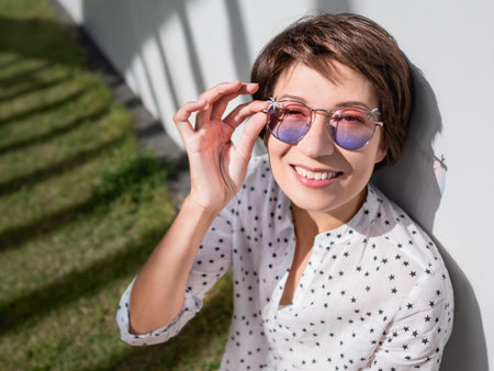 Smiling woman in colorful sunglasses has a rest on lawn in urban park. nature in town. Relax outdoors after work. summer vibes.の写真素材